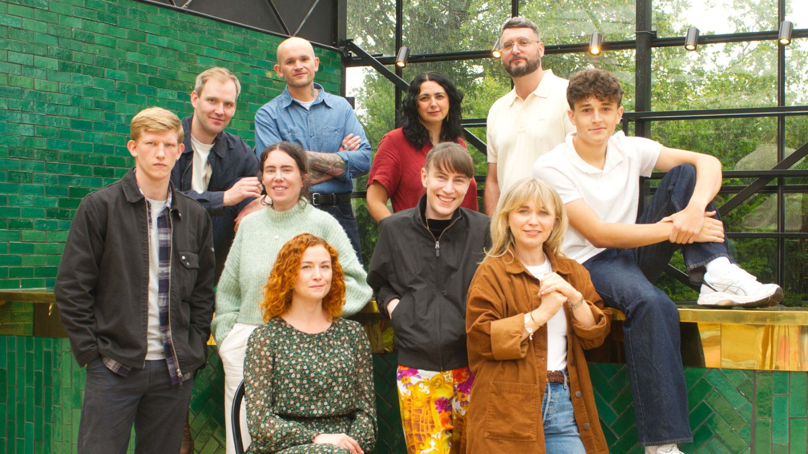 A medium outdoor group shot shows ten people posing in front of a green tiled bar with a glass and metal structure behind them.