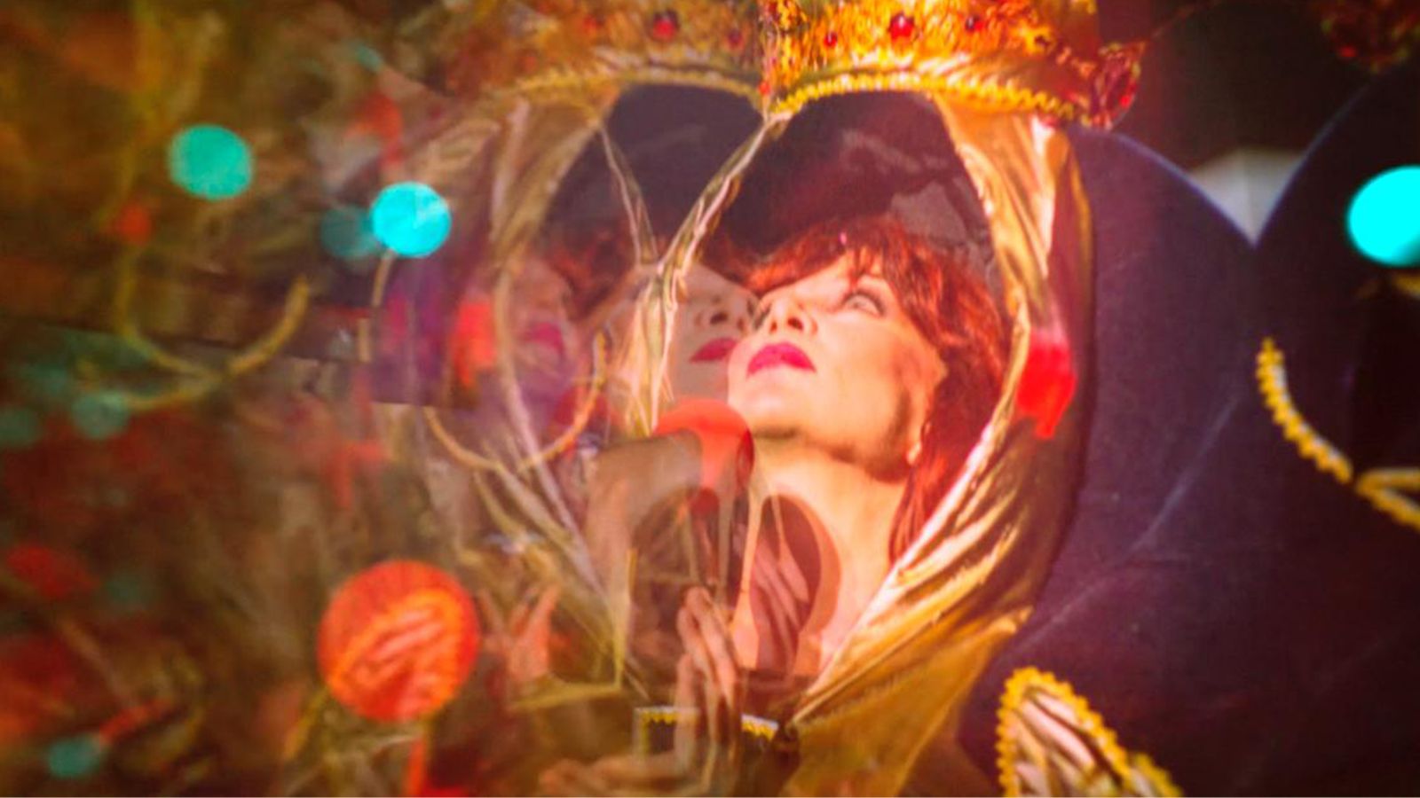 An artistic, multi-layered double exposure image depicting a woman with auburn hair and red lipstick looking upwards. Her face is framed by a golden, fabric-like border shaped like an arch or crown, adorned with small red gems. To the left, a mirrored, semi-transparent reflection of her face and hand is visible. Bright, out-of-focus light leaks in shades of teal and orange are scattered across the scene, creating a dreamlike, theatrical atmosphere.