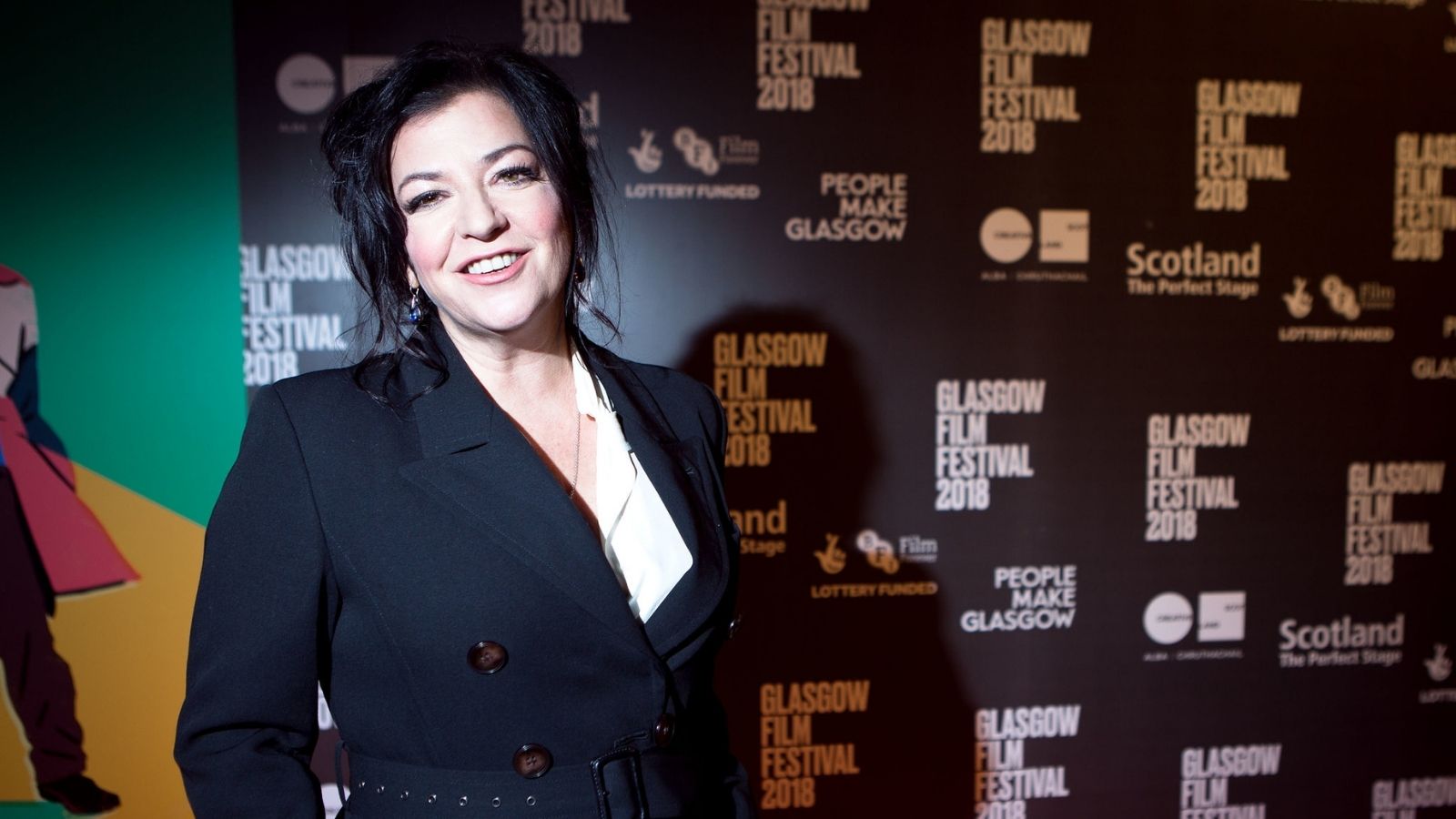 A woman with dark hair stands smiling at the Glasgow Film Festival 2018. She is wearing a black coat with a white inner top. The background features promotional signage for the festival.