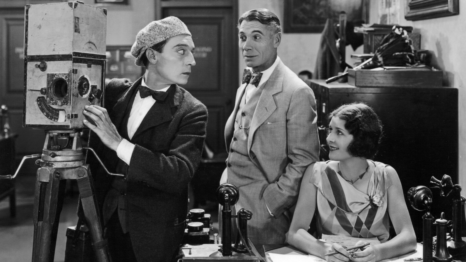 Black and white image of three people in a vintage office setting. A man in a suit and flat cap operates an old film camera on a tripod, looking to his left. Another man in a suit and bow tie stands watching him. A woman in a patterned dress sits at a desk with telephones, smiling towards the men.