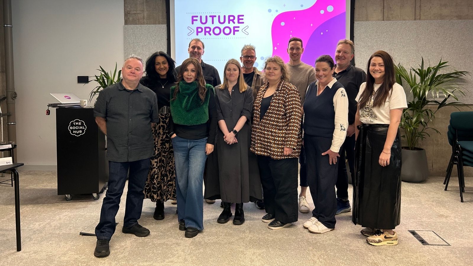 A group of eleven people standing indoors in front of a screen displaying 