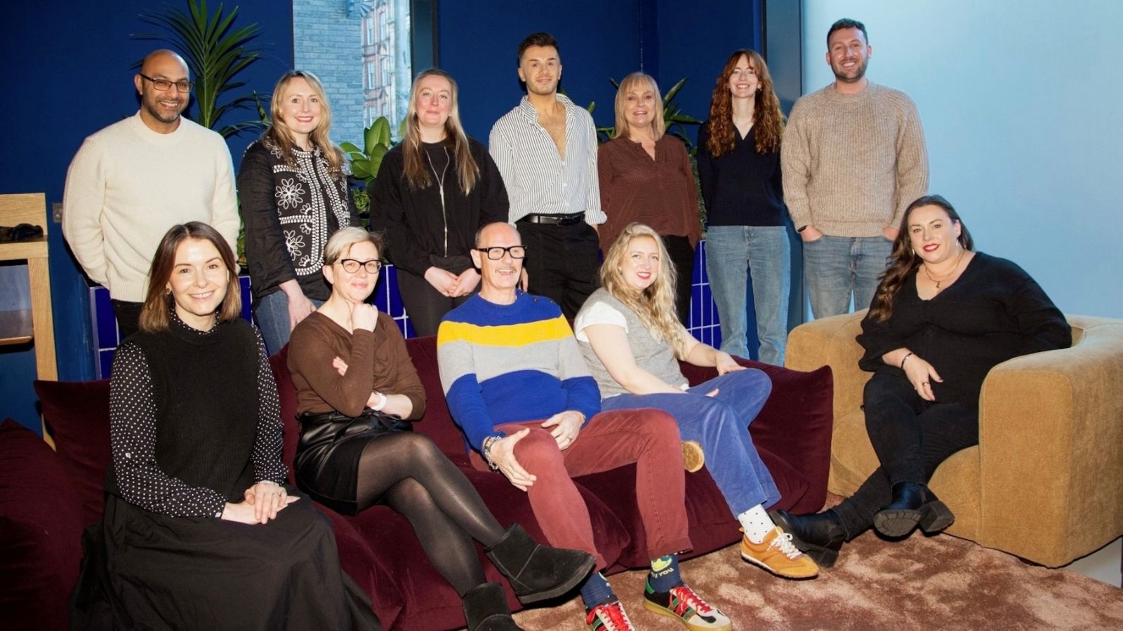 A group of twelve people is gathered in a cozy, brightly lit room with deep blue walls. They are casually dressed and positioned in two rows, with some sitting on a maroon sofa and others standing behind it. A large plant is visible in the background, and natural light filters through a window, enhancing the relaxed atmosphere.