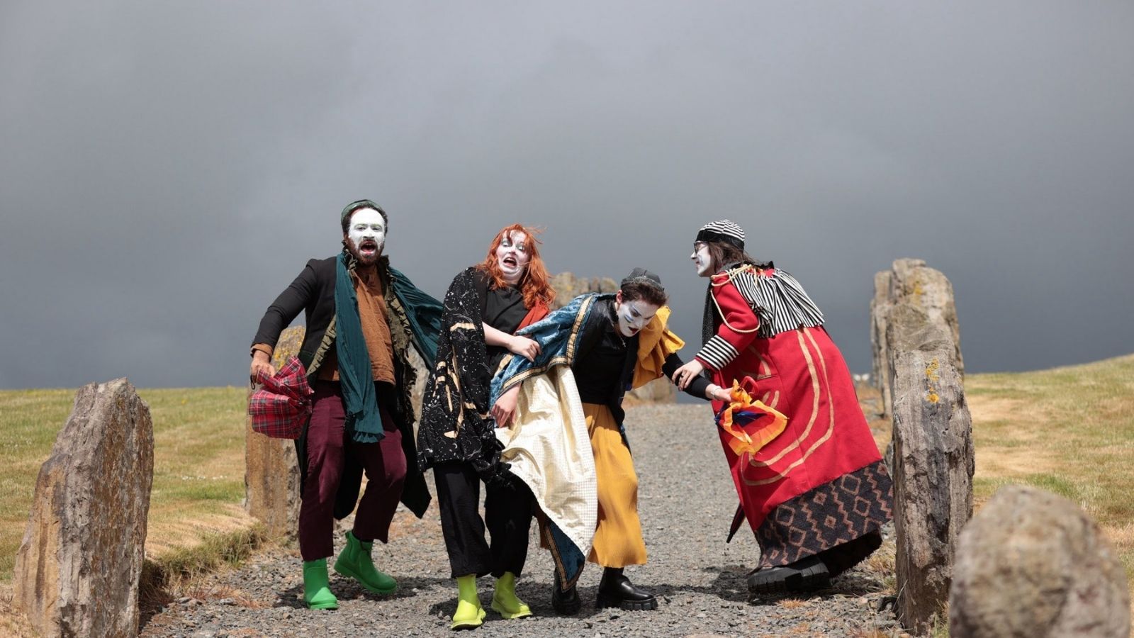 our people in theatrical costumes and white face paint stand on a gravel path between large stones. One wears green boots and holds a plaid bag, another in a flowing cape clings to the others with a dramatic expression. The sky is overcast, creating a dramatic backdrop.