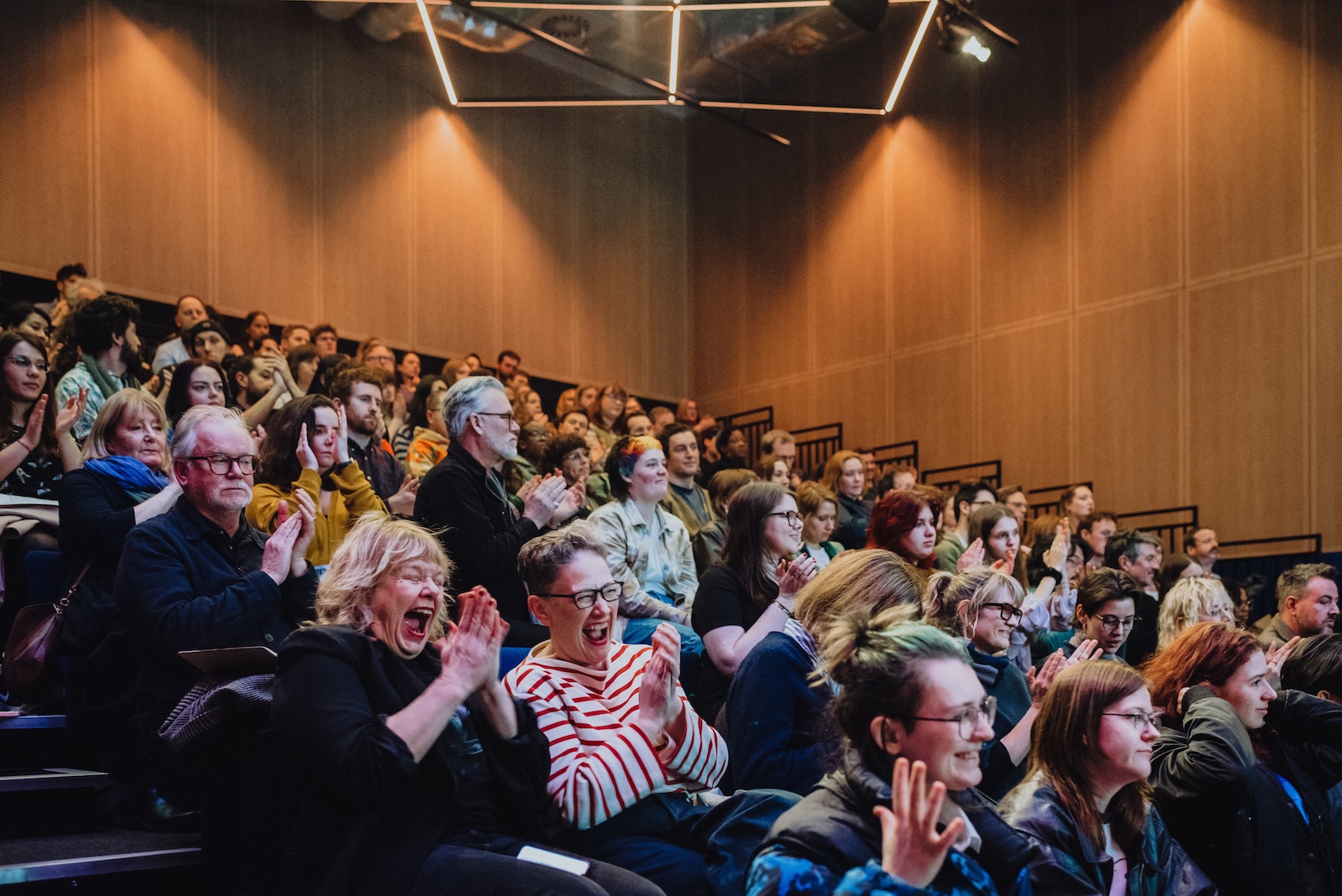 An professional audience of people of various ages in various poses of cheer, reacting to something amusing that has been said by the speaker on stage