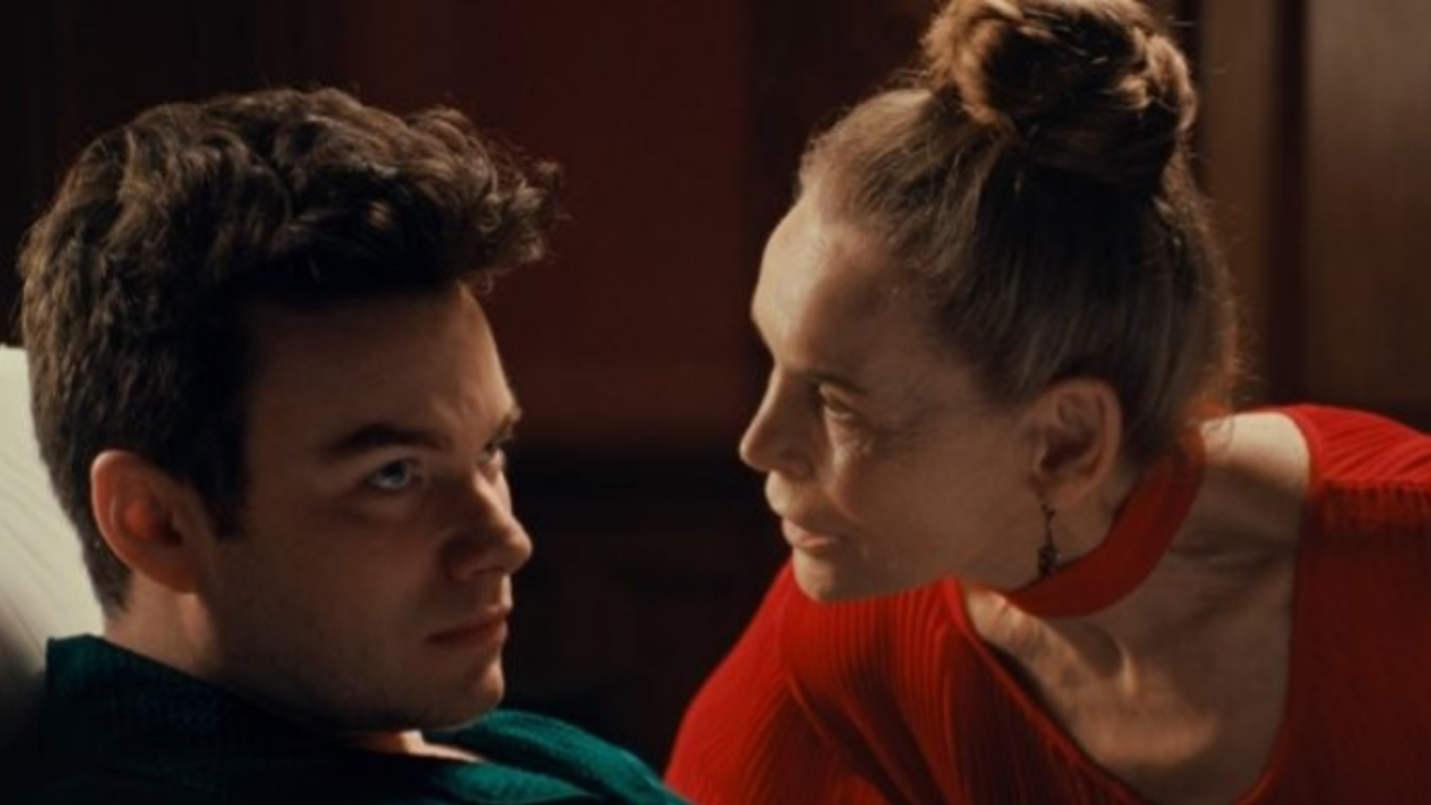 A woman in a bright red top intensely leans toward a man in a green sweater, who looks back at her with a serious expression. Both are indoors with dim lighting, creating a dramatic atmosphere.