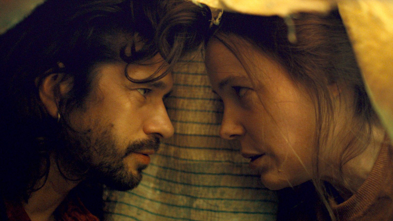 A man and woman gaze intently at each other, their foreheads almost touching. They are under a dimly lit, makeshift tent using striped fabric. The man's hair is long and curly, while the woman has her hair pulled back. The mood appears tense and intimate.
