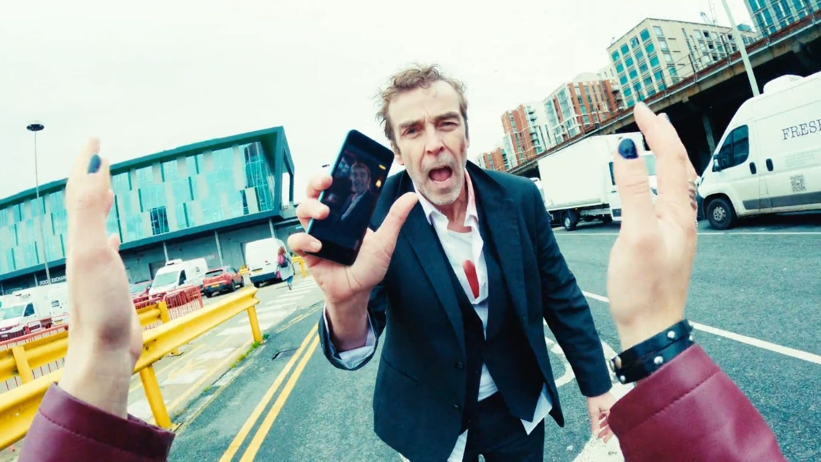 A man in a disheveled suit stands in the middle of the street, holding a smartphone with his image captured on its screen. His expression is anxious, with a partially open mouth. The scene includes a modern building, parked vehicles, and urban infrastructure in the background. Two hands, one wearing a bracelet, are raised in the frame, possibly reaching towards the man.