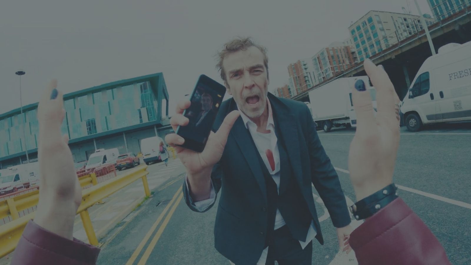 A man in a disheveled suit stands in the middle of the street, holding a smartphone with his image captured on its screen. His expression is anxious, with a partially open mouth. The scene includes a modern building, parked vehicles, and urban infrastructure in the background. Two hands, one wearing a bracelet, are raised in the frame, possibly reaching towards the man.