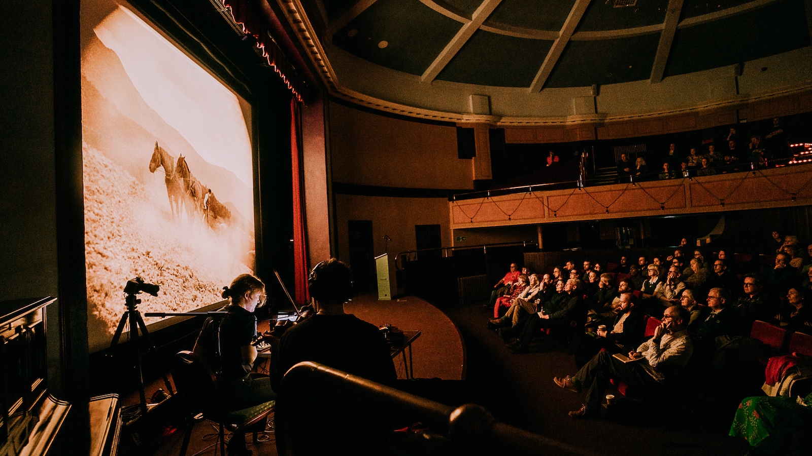 An audience in a dimly lit theater watches a large screen showing a sepia-toned image of two horses on a dusty landscape. A person operates equipment in the foreground, illuminated by the screen's glow. The theater has a vintage design with red curtains and balconies.
