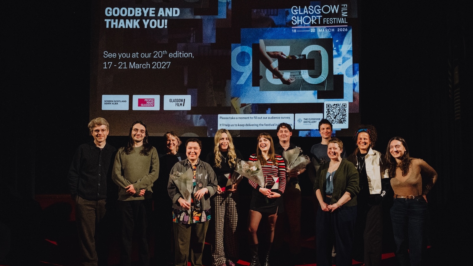 A group of smiling people standing in a row on a stage, holding bouquets of flowers. They are celebrating at the Glasgow Short Film Festival, with a large screen behind them displaying event details and sponsor logos. The screen includes text like 
