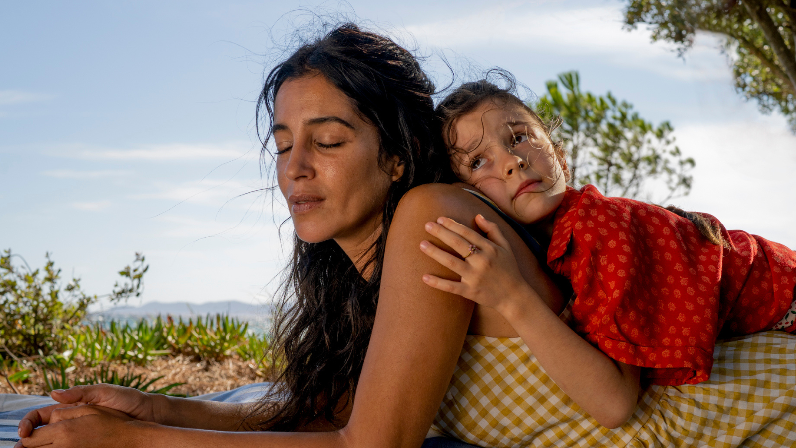 A medium close-up shows a woman with closed eyes lying on a blanket outdoors as a young girl in a red shirt rests on her back. The woman, with tan skin, dark, windblown hair, and a yellow gingham shirt, is in profile. The girl on her back, looking off to the right with a pensive expression, rests her chin on the woman’s shoulder and places one hand on her arm. Green foliage and a bright blue sky are visible in the background.