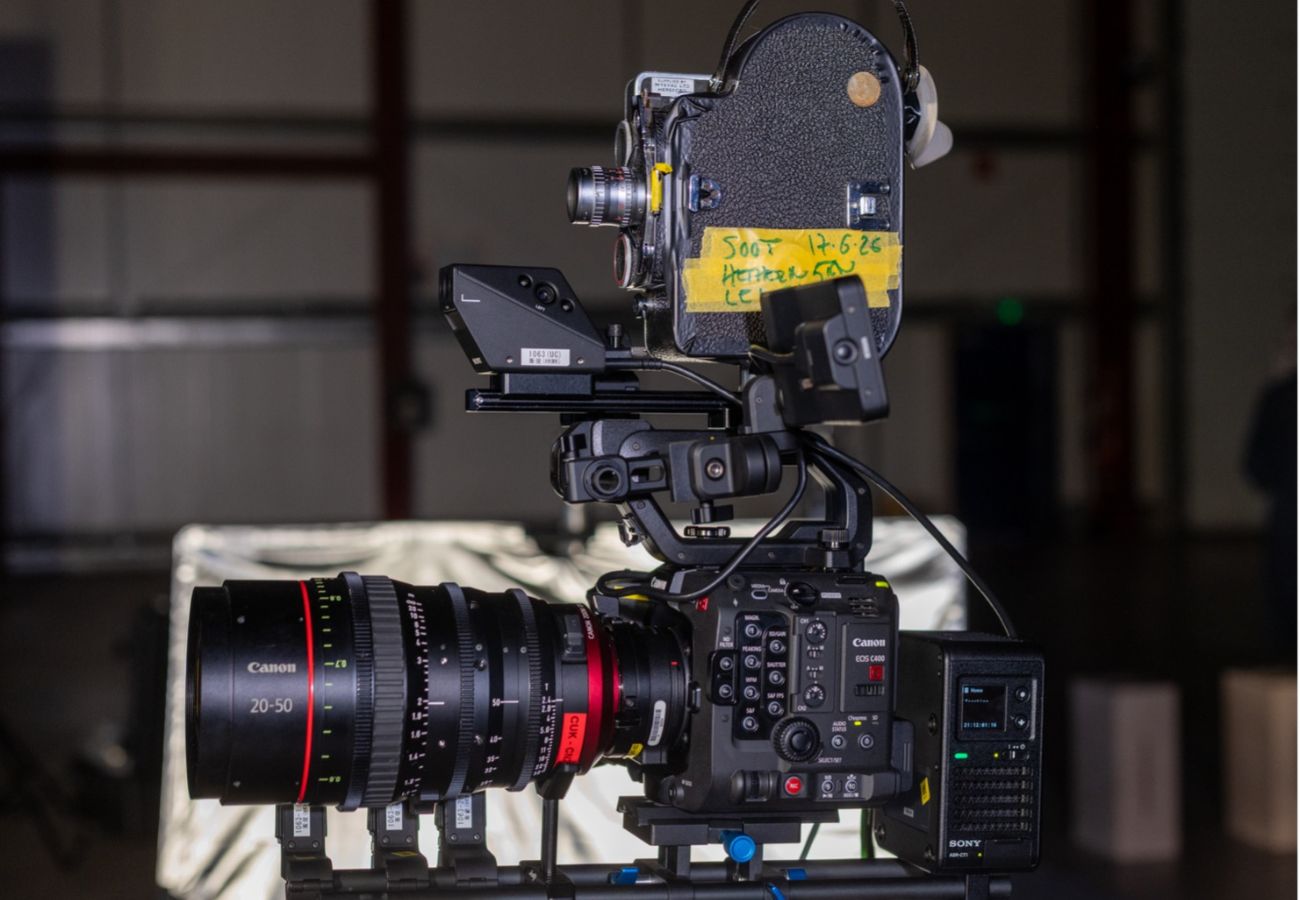 A professional Canon EOS C400 digital cinema camera is rigged on a rail system in profile, facing left, with a large Canon 20-50mm zoom lens. Mounted directly on top of the camera rig is a vintage, black textured movie camera with a piece of yellow tape that has handwritten notes. The setup is photographed in a dimly lit indoor space with a blurred background.