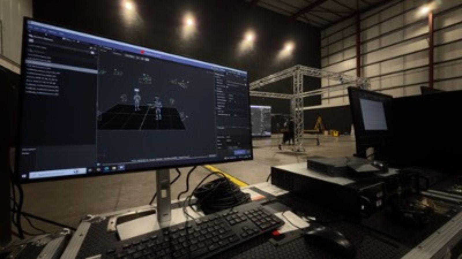 An eye-level, medium shot shows a computer monitor displaying a 3D animation interface with stick figures in a large, warehouse-style studio. In the foreground, a black keyboard, mouse, and other electronic equipment sit on a desk. Behind the primary monitor, another monitor is visible, and in the background, a large metal truss system stands in the centre of the warehouse. Overhead lights illuminate the spacious room.