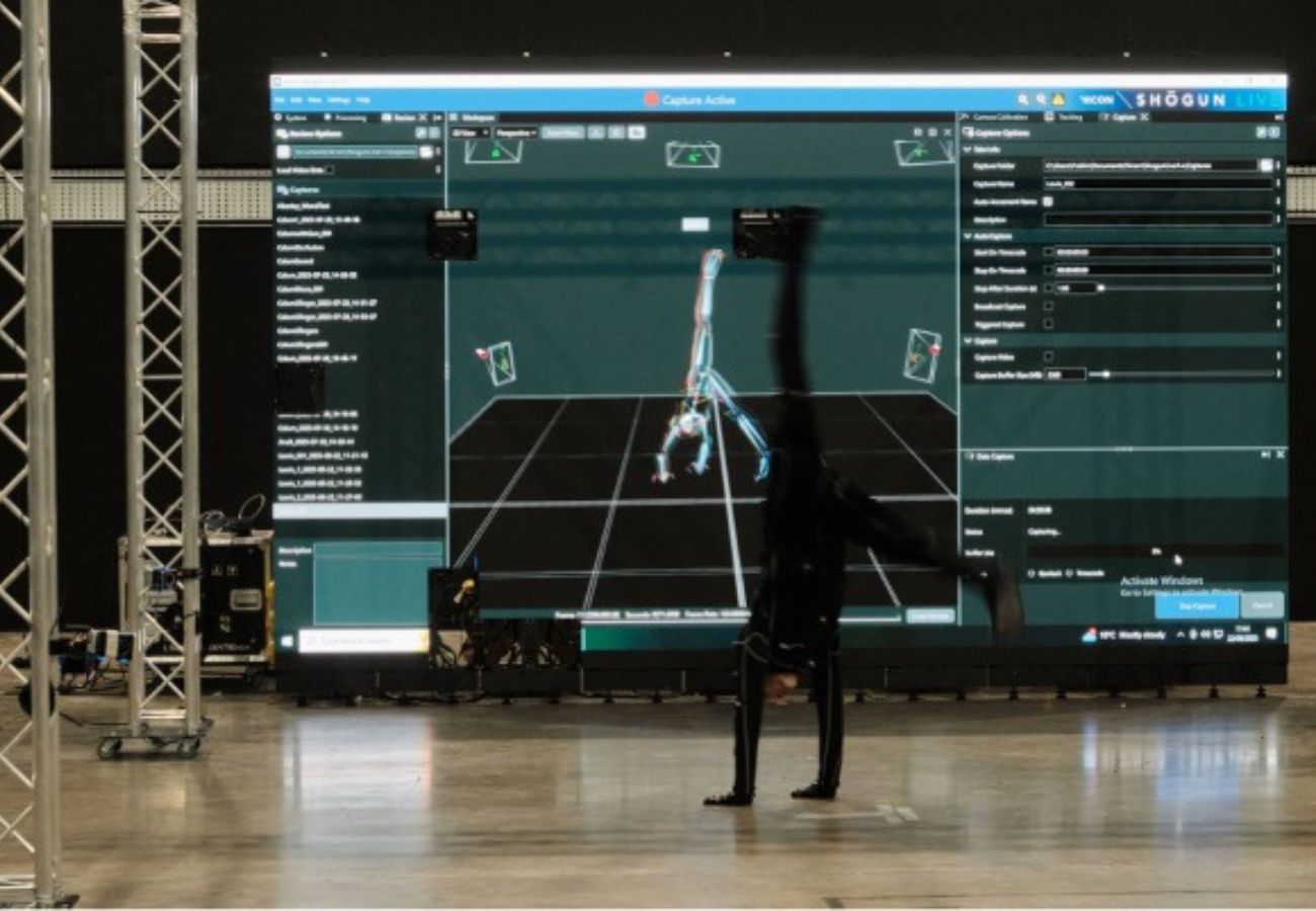 An actor clad in a black motion-capture suit performs a one-handed handstand in a studio. In the background, a large digital screen displays the Shogun Live software interface, showing a real-time 3D wireframe skeleton mirroring the performer's acrobatic pose on a virtual grid. Metal truss structures stand to the left on a polished concrete floor.