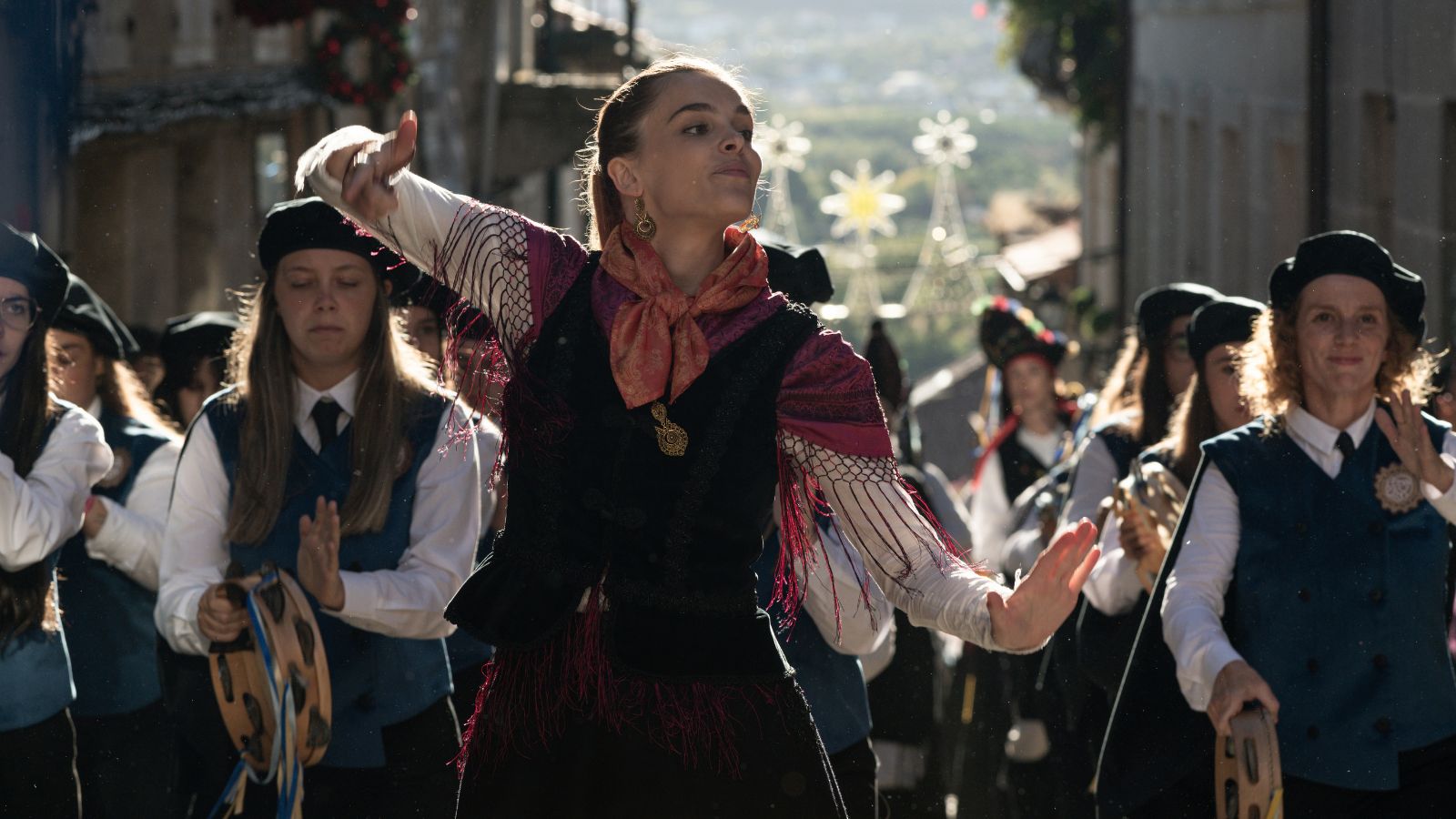 A close-up shot captures a young woman dancing in a traditional parade. She wears a black vest over a white long-sleeved shirt, decorated with a red, patterned scarf tied around her neck and dark-red fringed accents on her sleeves. Behind her, a group of people wearing matching dark blue vests and black caps look on, some playing traditional hand drums. The background is a cobblestone street decorated with illuminated star-shaped holiday lights.