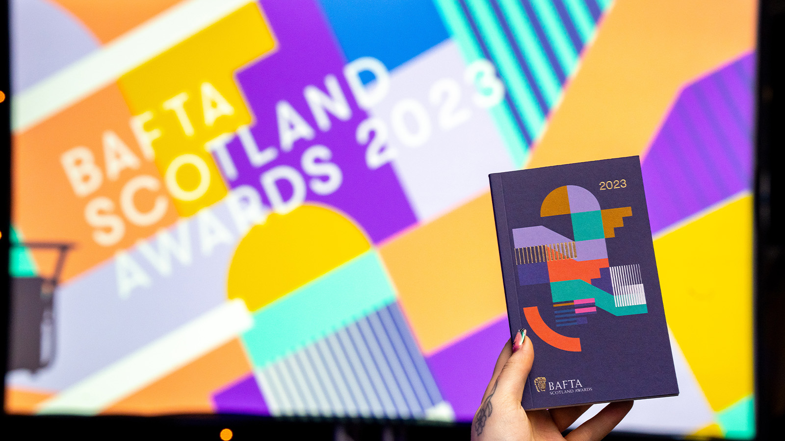 A hand holds up the print version of the BAFTA Scotland Awards 2023 programme in front of a general view of preparations ahead of the 2023 BAFTA Scotland Awards held at the DoubleTree by Hilton Glasgow Central on November 19, 2023 in Glasgow, Scotland. (Photo by John Clark/BAFTA via Getty Images)