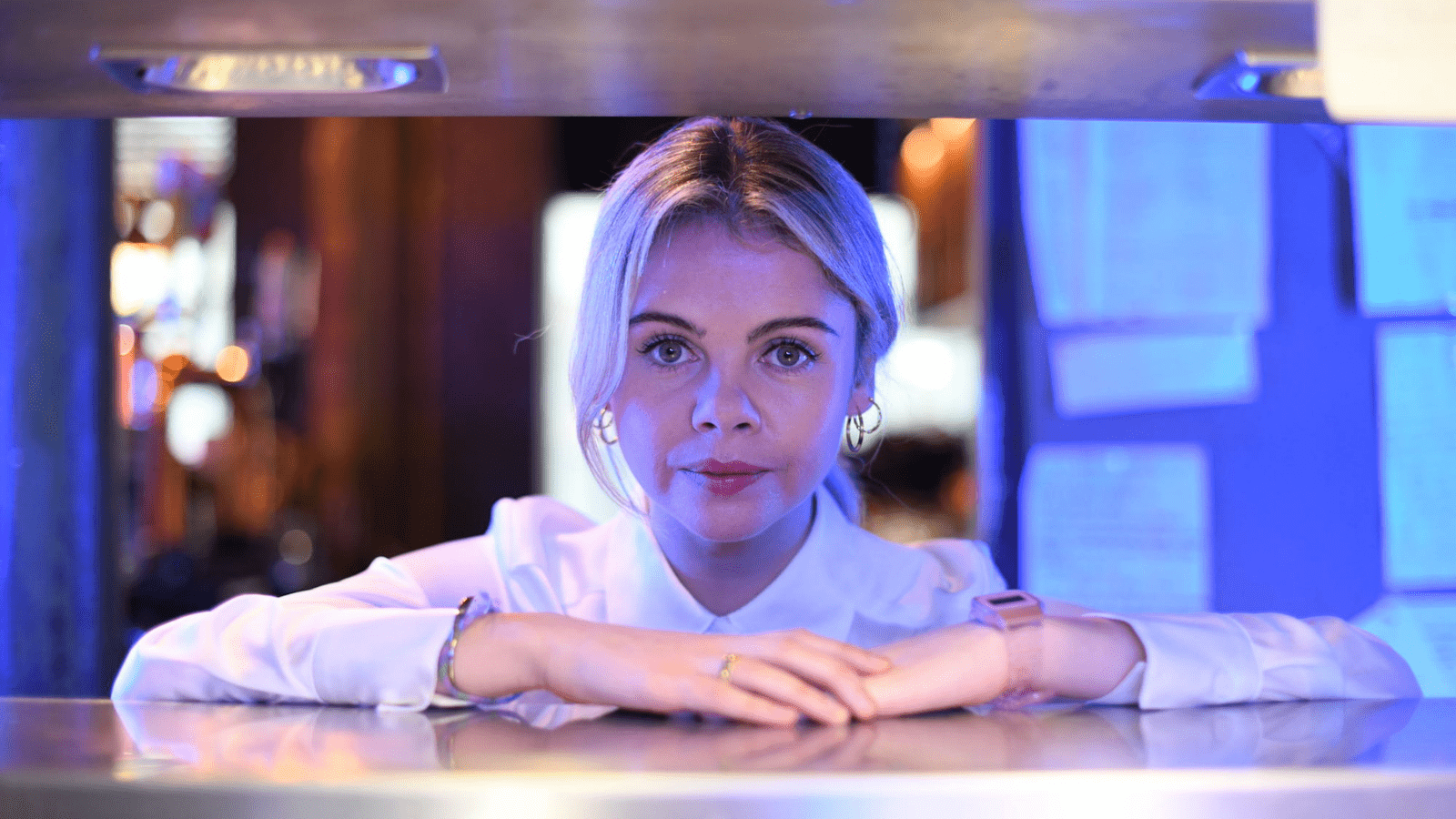 Blonde woman in white shirt stares at the camera while leaning against a counter.