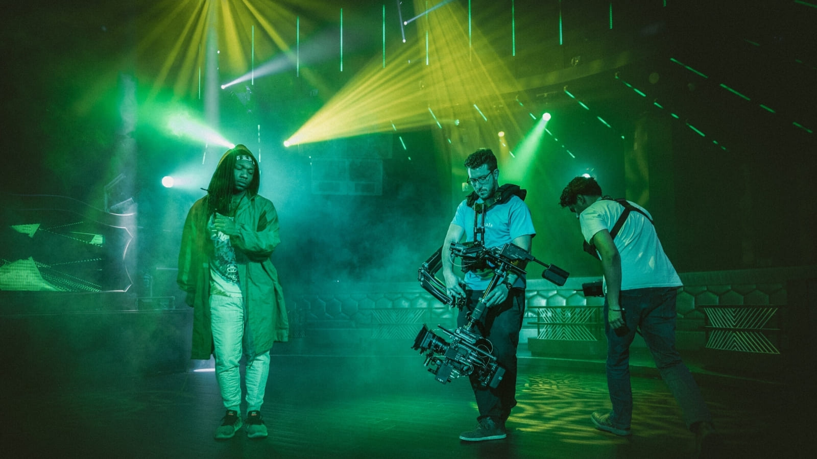 A camera operator and a member of film crew stand next to a performer in an arena style warehouse, filming what looks like a misty music video. The scene is tinged with green light