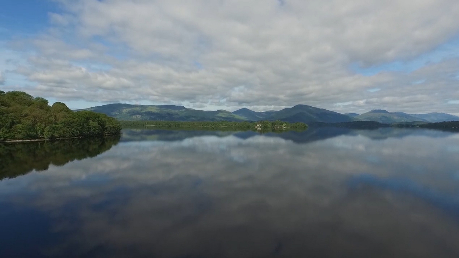 A loch in front of a wide area of green trees under a blue sky