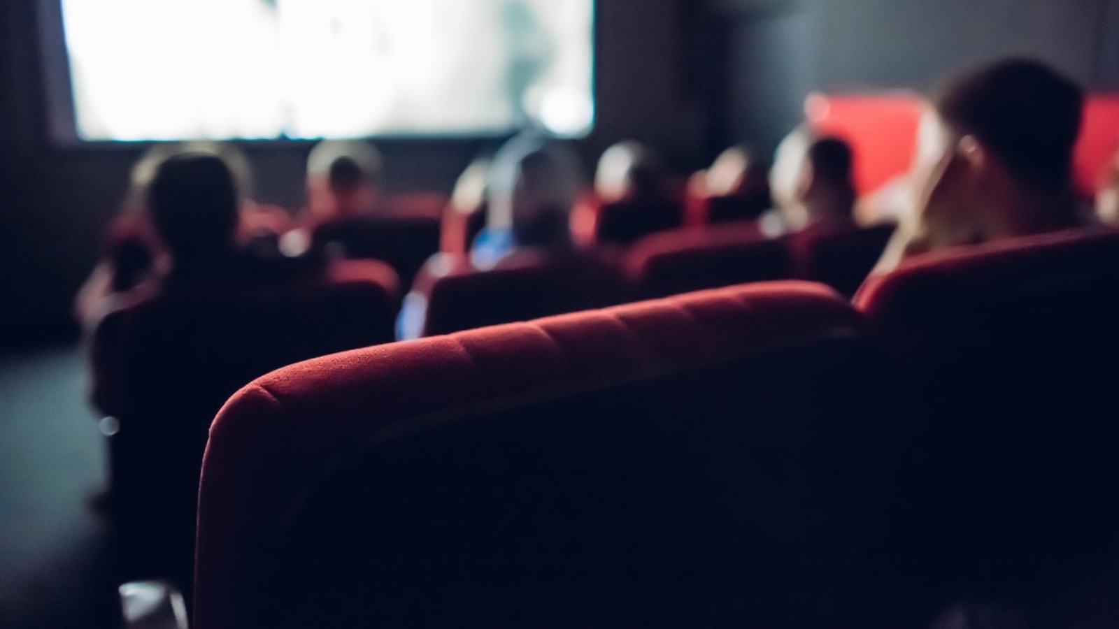 Close up of red cinema seats in a dark auditorium, some people sit on chairs in the distance, watching the screen