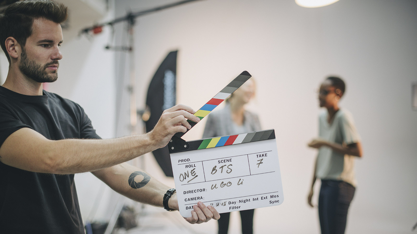 A man holds a clapper board on a set, two people appear blurred in the background