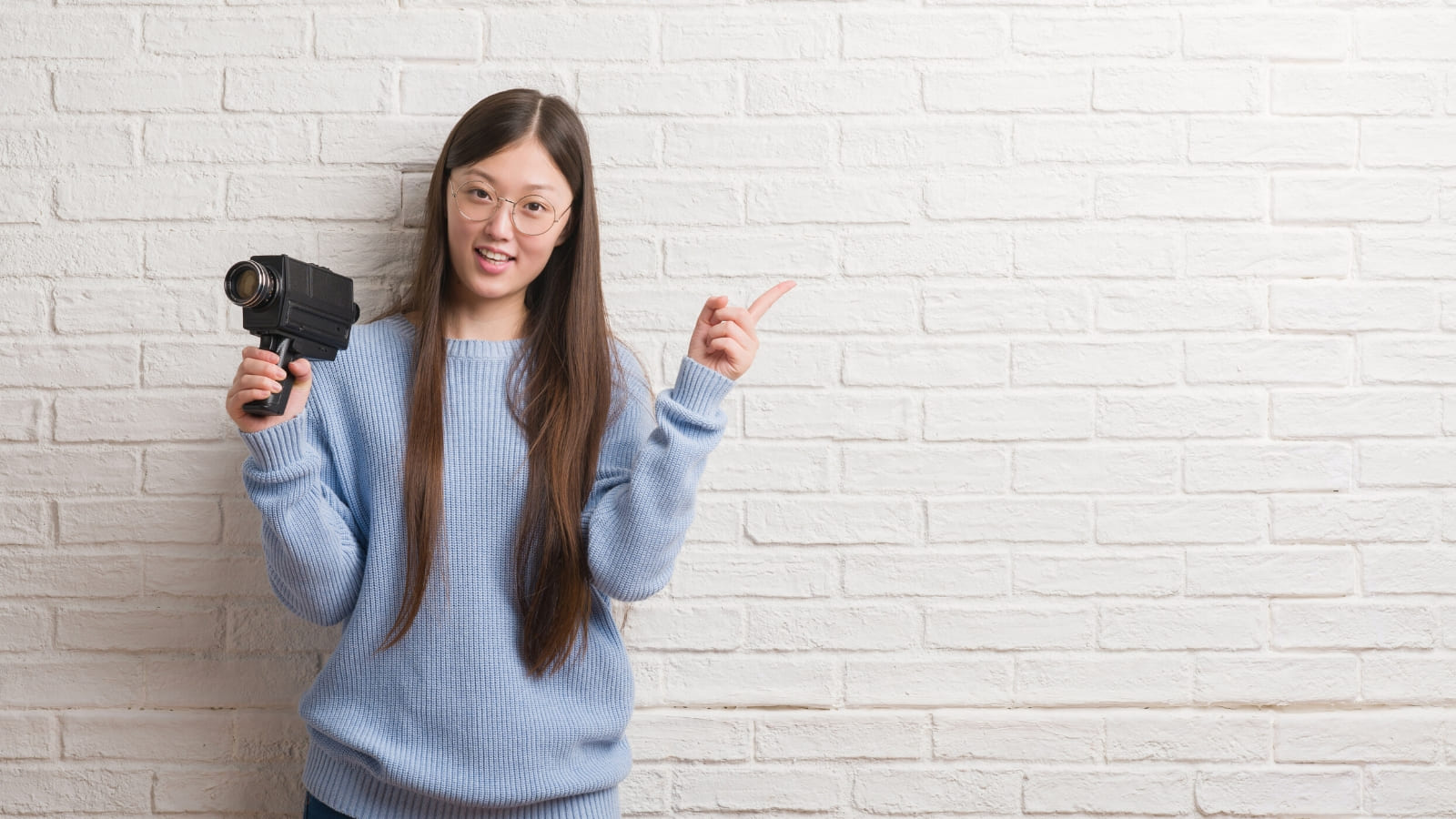 A young person holds a film camera whilst pointing their finger, standing against a white wall backdrop