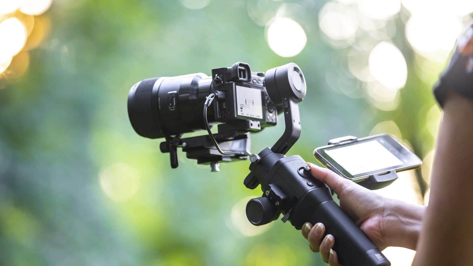 A hand holds up a camera on a gimbal device, pointing in front of a green background of nature