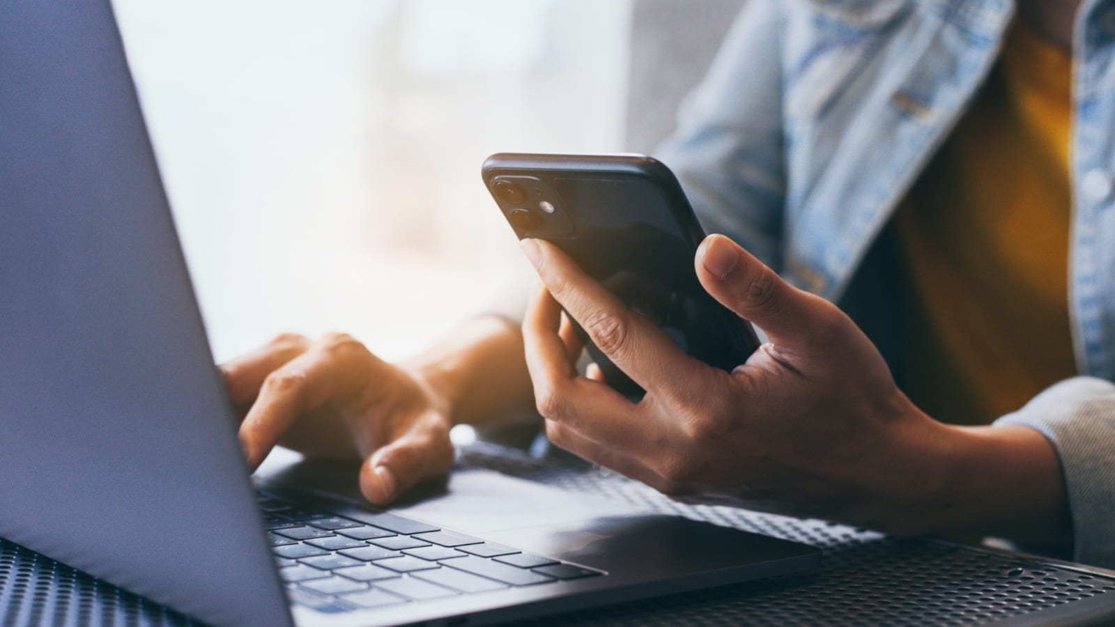 A person sits typing on their laptop while looking at their phone