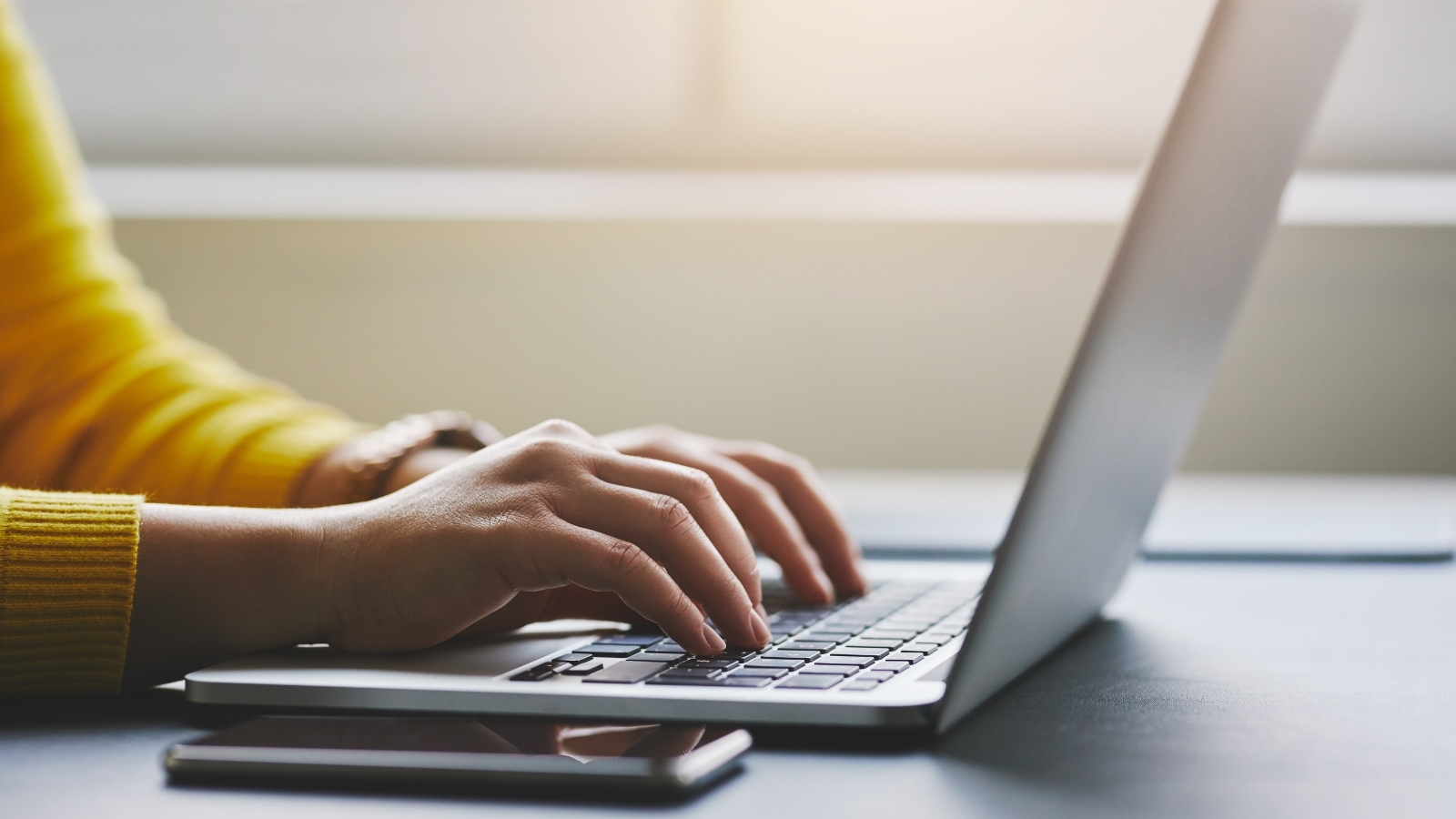 A person sits at a laptop typing, a phone lies on the desk next to the laptop