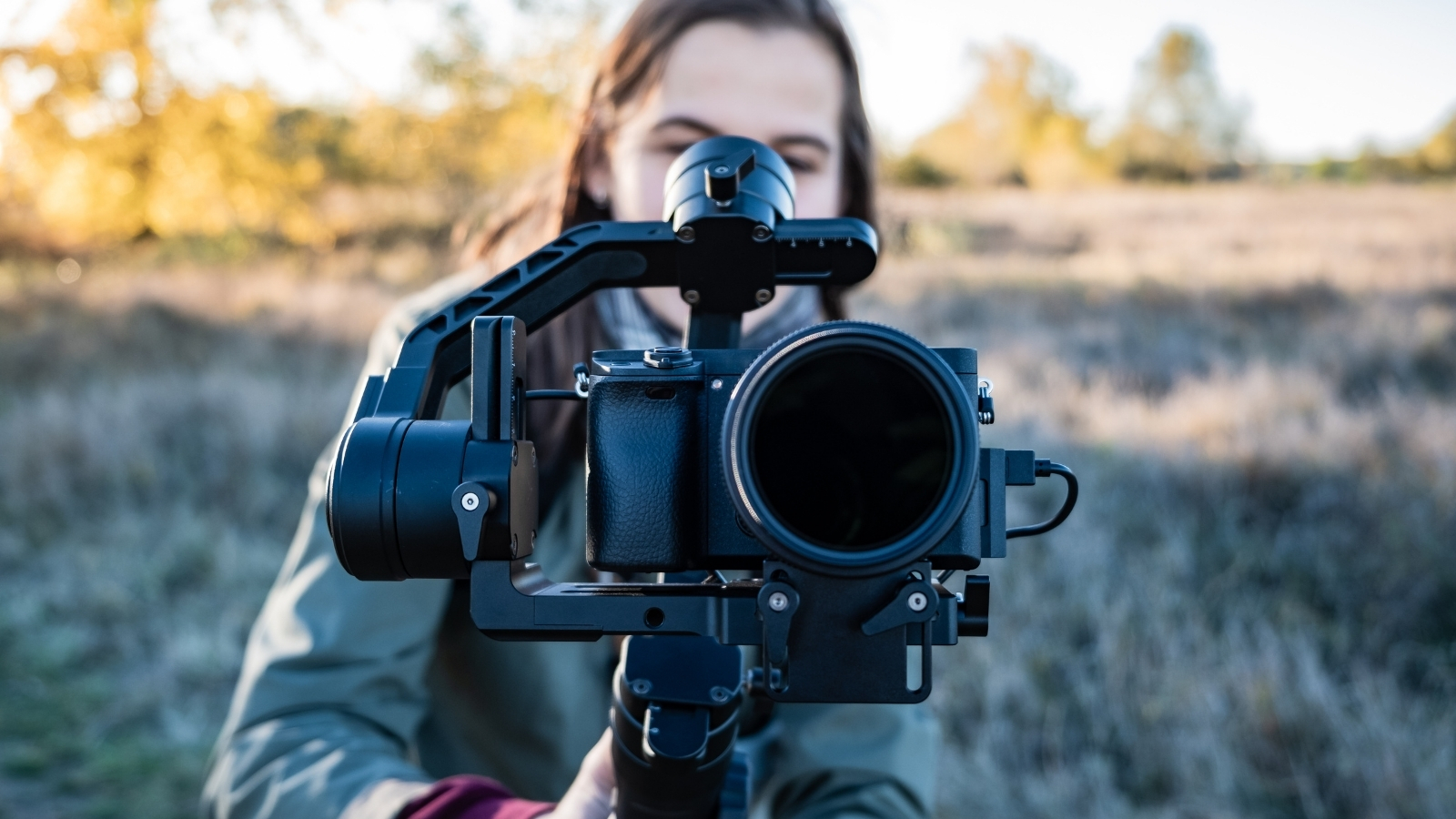 A person stands in a chilly field with a tree in the background, holding up a DSLR camera inside a handheld gimbal stabiliser