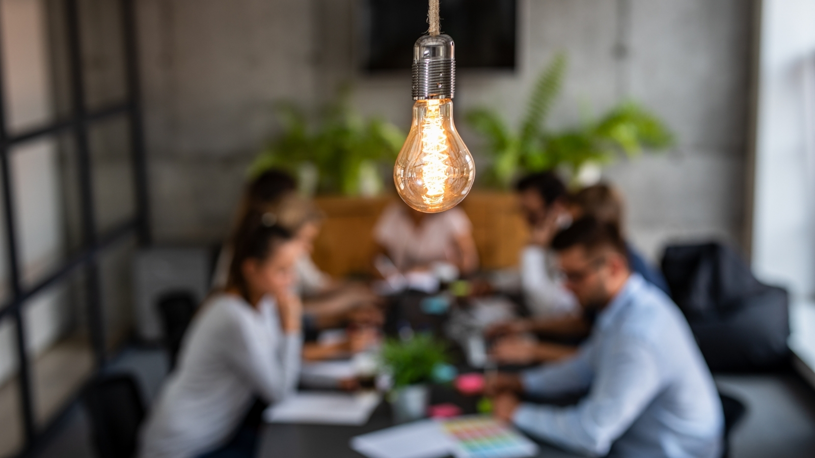 A group of creative producers collaborate at an office desk, they are slightly blurred out of the image, with a big lightbulb in focus hanging above them